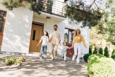 A happy family of four — two parents and two young children — holding hands and walking together along a paved path in front of a modern white home